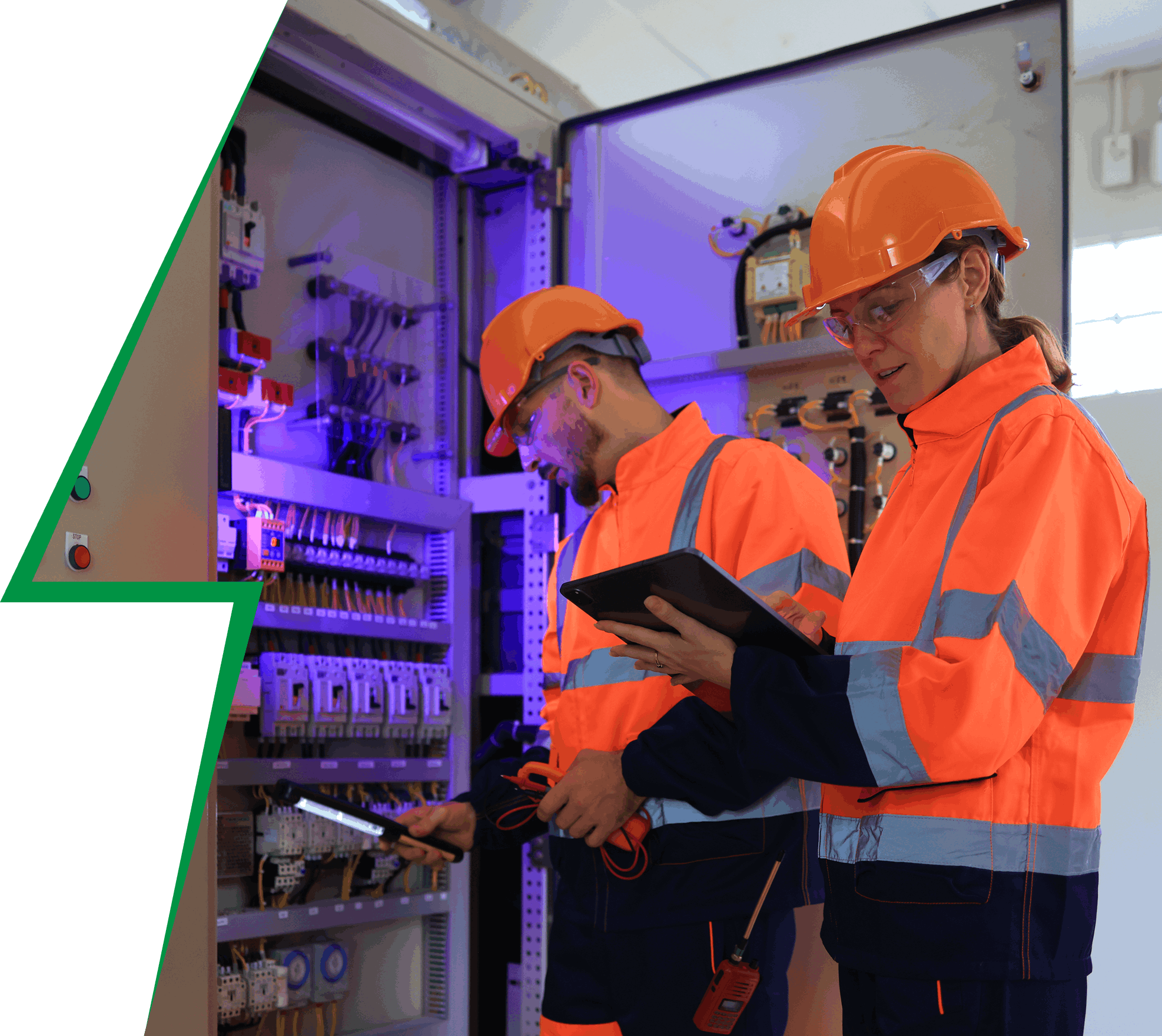 Two workers in safety gear inspect an electrical panel. One holds a tablet, the other uses a tool. Both wear orange helmets and reflective jackets.