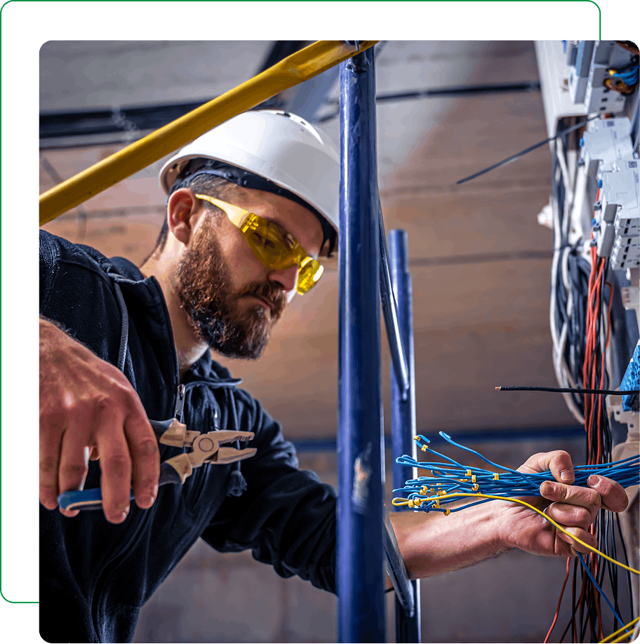 A man wearing a hard hat and safety glasses is using pliers to work with electrical wires in an industrial setting.